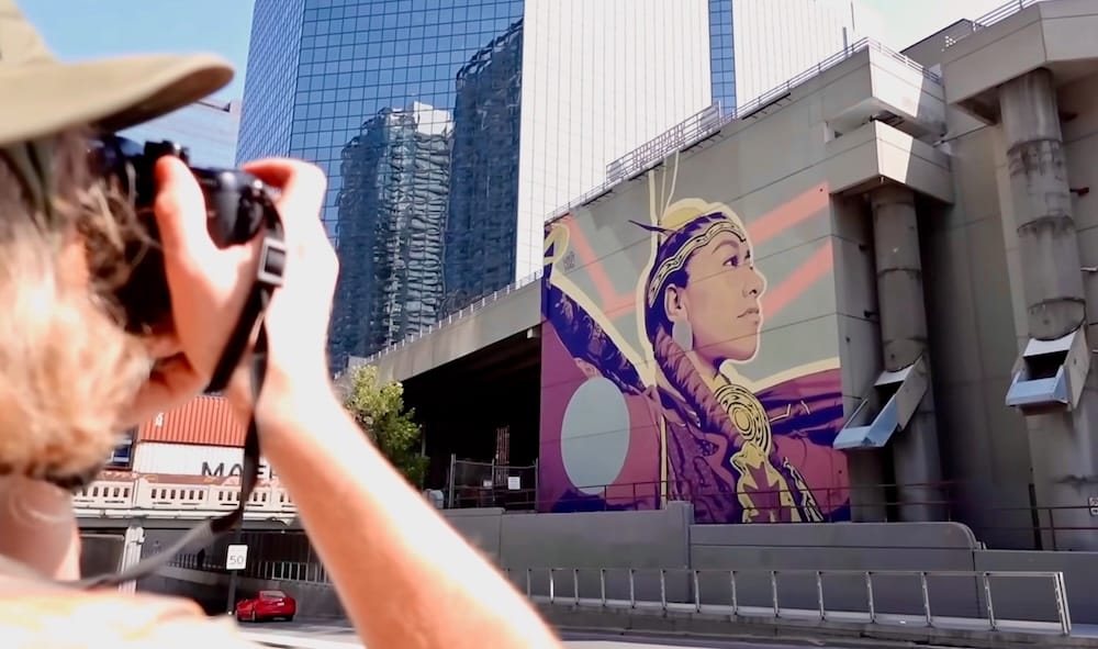 Samuel Jeffery photographs a large mural of an Indigenous woman on a downtown Calgary building in Calgary, Alberta, Canada, with glass office towers and elevated concrete structures framing the street art scene.