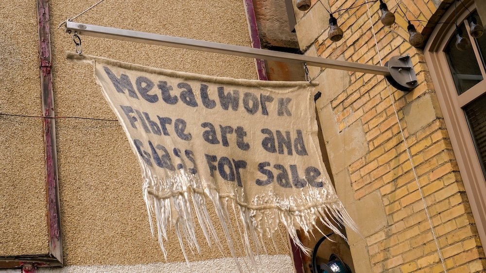 Handmade art sign in downtown Fernie Hand-painted fabric sign reading “metalwork, fibre art and glass for sale” hanging from a brick building in downtown Fernie, British Columbia, capturing the town’s creative small-business spirit and walkable heritage streetscape.