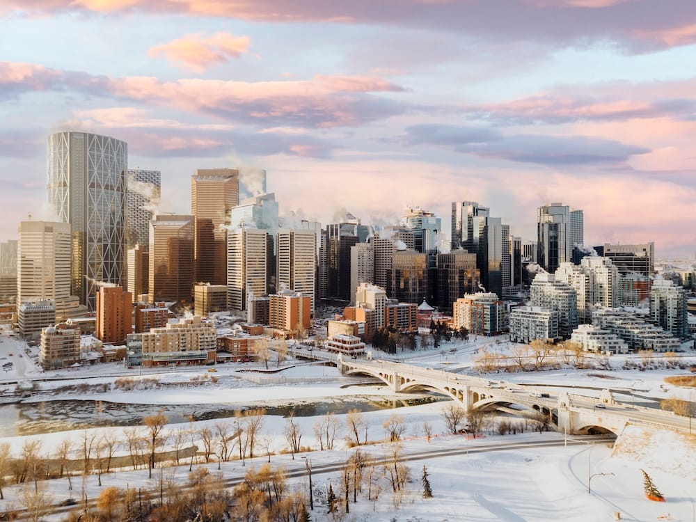 Downtown Calgary's skyline dusted with snow at sunset, with the frozen Bow River, a bridge, and steam rising from buildings against a pink winter sky, Alberta, Canada