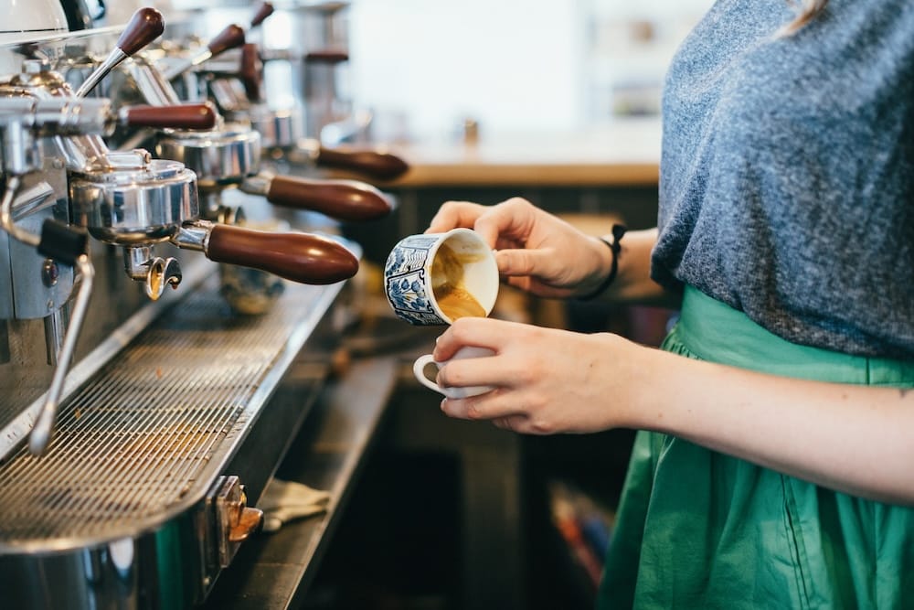 Una barista prepara café en una máquina de espresso en Edmonton, Alberta, Canadá, vertiendo desde una pequeña taza decorada hacia otra junto a los cabezales metálicos de la máquina.