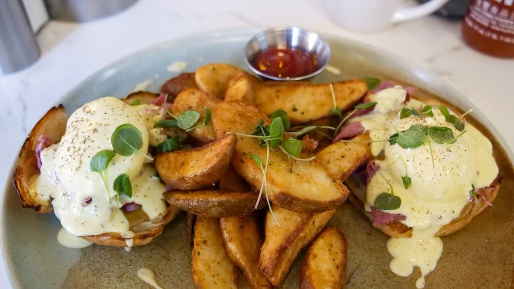 Close-up of an Edmonton, Alberta, Canada brunch plate with two eggs Benedict covered in hollandaise, roasted potato wedges, microgreens and a small dish of sauce on the side.