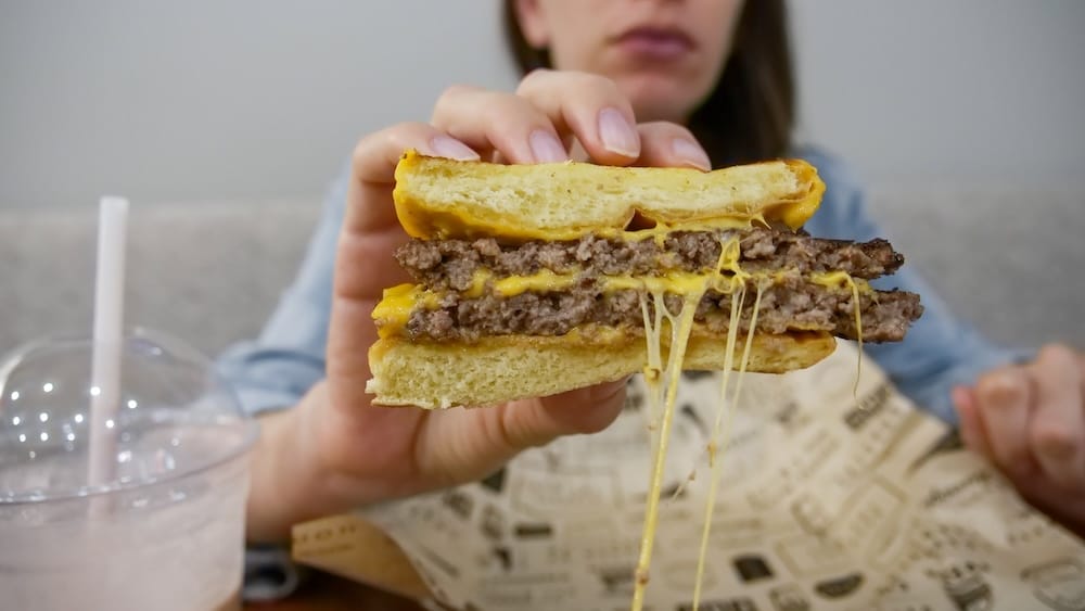 Queso estirándose de la hamburguesa con batido, Wayback Burgers, Edmonton, Alberta, Canadá — Audrey Bergner Audrey Bergner sostiene la mitad de una hamburguesa con queso con queso derretido estirándose en Wayback Burgers en Edmonton, Alberta, Canadá, con un batido y una canasta forrada con papel visibles en la mesa frente a ella.