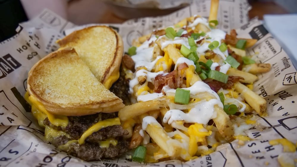 Canasta con hamburguesa doble con queso y papas fritas cargadas, Wayback Burgers, Edmonton, Alberta, Canadá Una hamburguesa doble con queso en un pan tostado y mantecoso servida junto a papas fritas cargadas con queso y panceta en una canasta forrada con papel de la marca en Wayback Burgers en Edmonton, Alberta, Canadá.