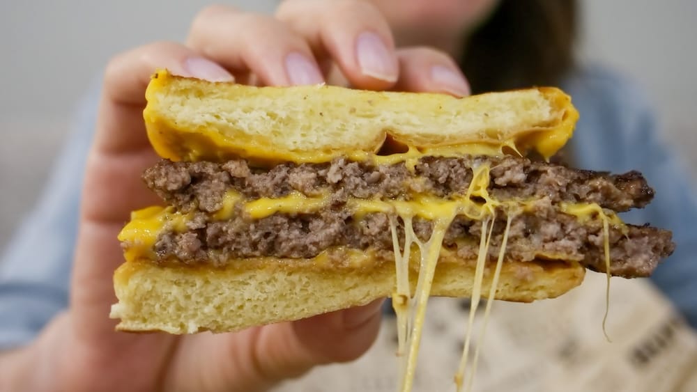 Hamburguesa doble con queso derretido estirándose, Wayback Burgers, Edmonton, Alberta, Canadá — Audrey Bergner Audrey Bergner sostiene la mitad de una hamburguesa doble con queso de Wayback Burgers en Edmonton, Alberta, Canadá, mostrando dos medallones de carne, queso derretido estirándose entre las capas y un pan dorado y mantecoso.