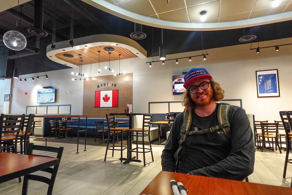 Interior del restaurante Wayback Burgers, Edmonton, Alberta, Canadá — Samuel Jeffery Samuel Jeffery sentado adentro de Wayback Burgers en Edmonton, Alberta, Canadá, usando una gorra de Edmonton y una mochila, con asientos tipo box, televisores en la pared y un cartel con la bandera de Canadá visible de fondo.