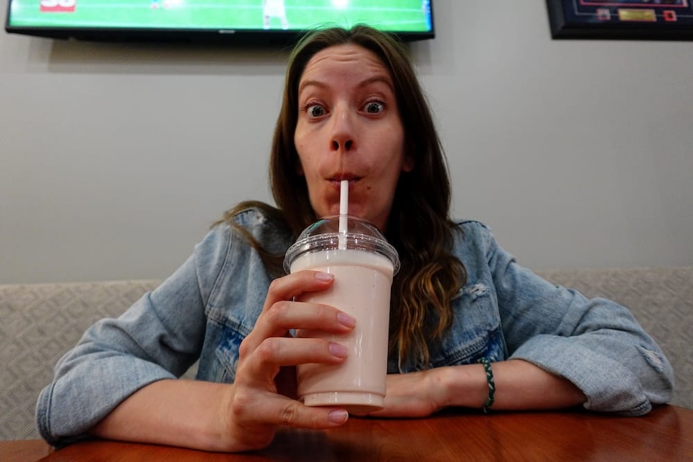 Batido de frutilla en Wayback Burgers, Edmonton, Alberta, Canadá — Audrey Bergner Audrey Bergner toma un batido de frutilla con un sorbete en Wayback Burgers en Edmonton, Alberta, Canadá, haciendo una expresión juguetona con los ojos muy abiertos mientras está sentada en un box del restaurante.