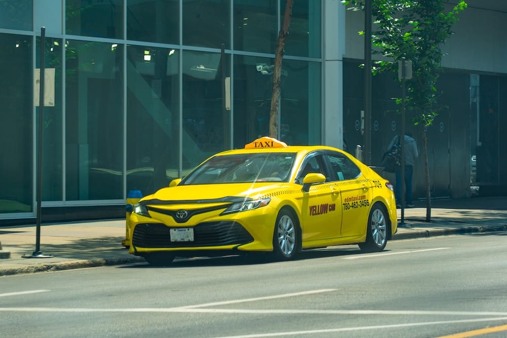 Taxi amarillo en el centro de Edmonton, Alberta, Canadá, para traslados urbanos y desde el aeropuerto Un taxi amarillo de Edmonton estacionado en una calle del centro de la ciudad, Alberta, Canadá, con edificios de vidrio y árboles bordeando la vereda detrás del vehículo.