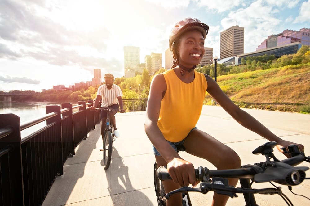 Edmonton bike tour through the river valley with downtown skyline views, Alberta, Canada Two cyclists ride along a paved path in Edmonton’s river valley, Alberta, Canada, with the North Saskatchewan River beside them and the downtown skyline rising in the background.