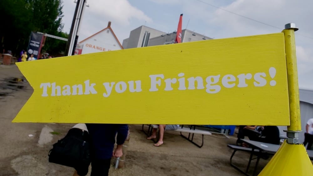 Bright yellow “Thank you Fringers!” sign at the Edmonton Fringe Festival in Old Strathcona, Edmonton, Alberta, Canada, with festivalgoers, benches and venue buildings behind.