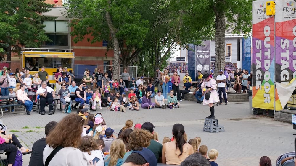 Un artista callejero disfrazado entretiene a un gran público sentado en el escenario al aire libre (Outdoor Stage) del Edmonton International Fringe Theatre Festival en Old Strathcona, Edmonton, Alberta, Canadá, con banners del festival y un food truck de fondo.