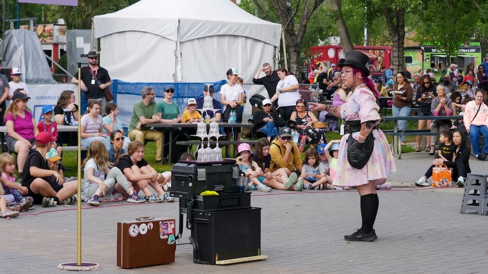 Artista Callejera en el Predio del Edmonton Fringe Festival, Old Strathcona, Edmonton, Alberta, Canadá Una artista callejera con galera y vestuario en tonos pastel entretiene a un público sentado al aire libre en el predio del Edmonton Fringe Festival, en Old Strathcona, Edmonton, Alberta, Canadá, con food trucks y carpas de fondo.