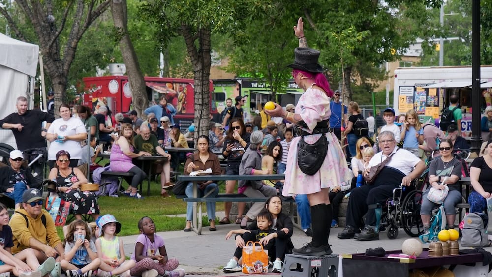 Edmonton Fringe street performance with audience in Old Strathcona, Edmonton, Alberta, Canada Costumed Edmonton Fringe street performer entertaining a seated crowd beneath trees, with food trucks, picnic tables and festival activity in Old Strathcona, Edmonton, Alberta, Canada.