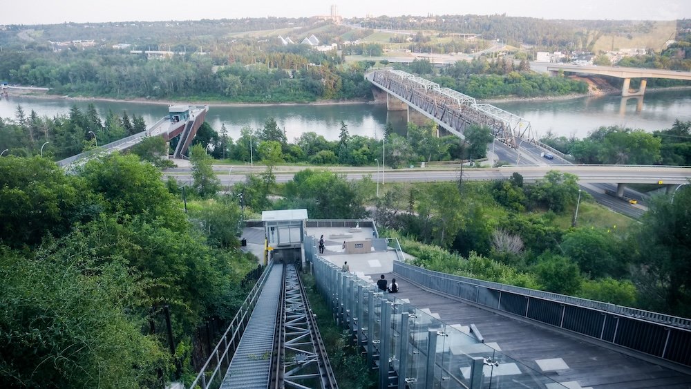 Edmonton funicular descending into the North Saskatchewan River valley. View down the Edmonton funicular descending from the hotel into the North Saskatchewan River valley, with glass railings, forested slopes, a pedestrian bridge, highway overpasses and the wide river framed by greenery at sunset.