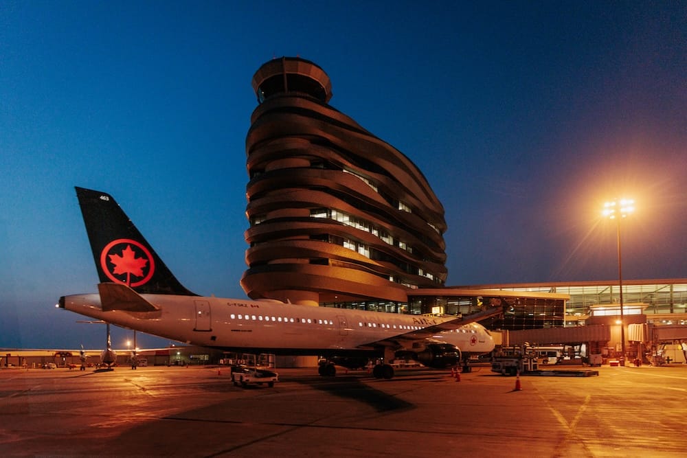 Terminal de YEG y avión de Air Canada de noche en el Aeropuerto Internacional de Edmonton, Alberta, Canadá Un avión de Air Canada estacionado junto a la terminal iluminada del Aeropuerto Internacional de Edmonton, Alberta, Canadá, con la torre del aeropuerto bajo un cielo azul profundo al anochecer.