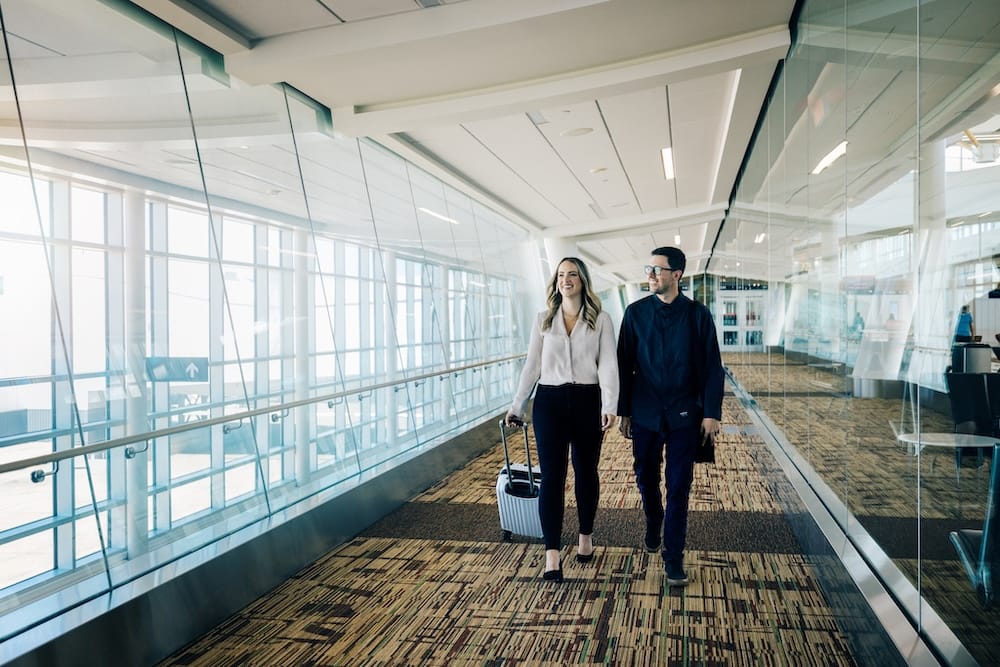 Pareja caminando con equipaje dentro del Aeropuerto Internacional de Edmonton YEG, Alberta, Canadá Una pareja camina por un pasillo luminoso con paredes de vidrio dentro del Aeropuerto Internacional de Edmonton, Alberta, Canadá, mientras una de las personas lleva una valija de mano con ruedas.