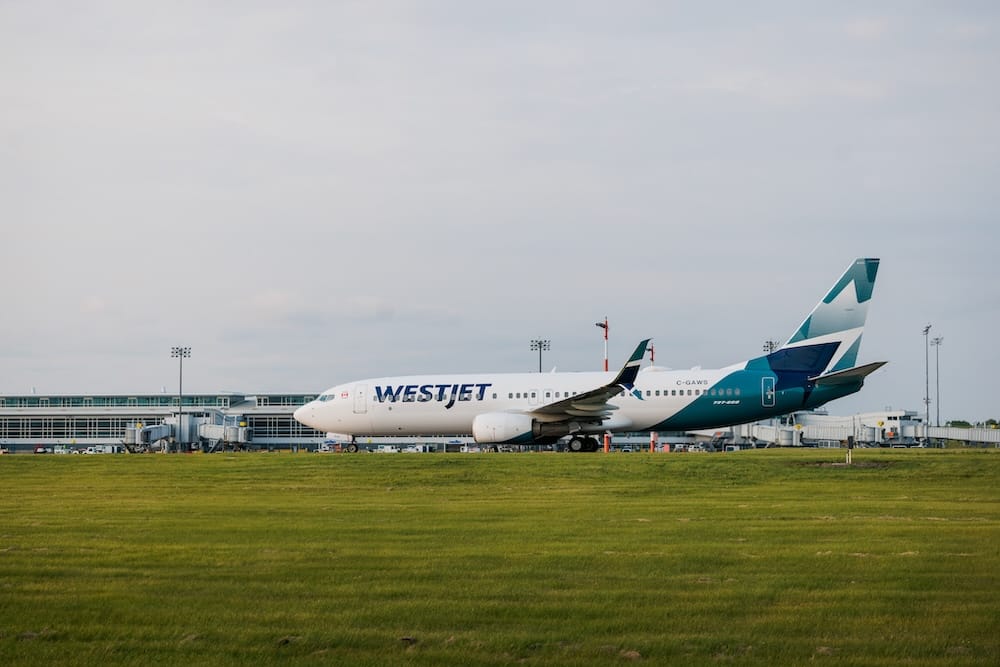 Avión de WestJet junto a la terminal del Aeropuerto Internacional de Edmonton YEG, Alberta, Canadá Un Boeing 737 de WestJet está junto a la terminal del Aeropuerto Internacional de Edmonton, Alberta, Canadá, con una amplia extensión de césped en primer plano bajo un cielo nublado.