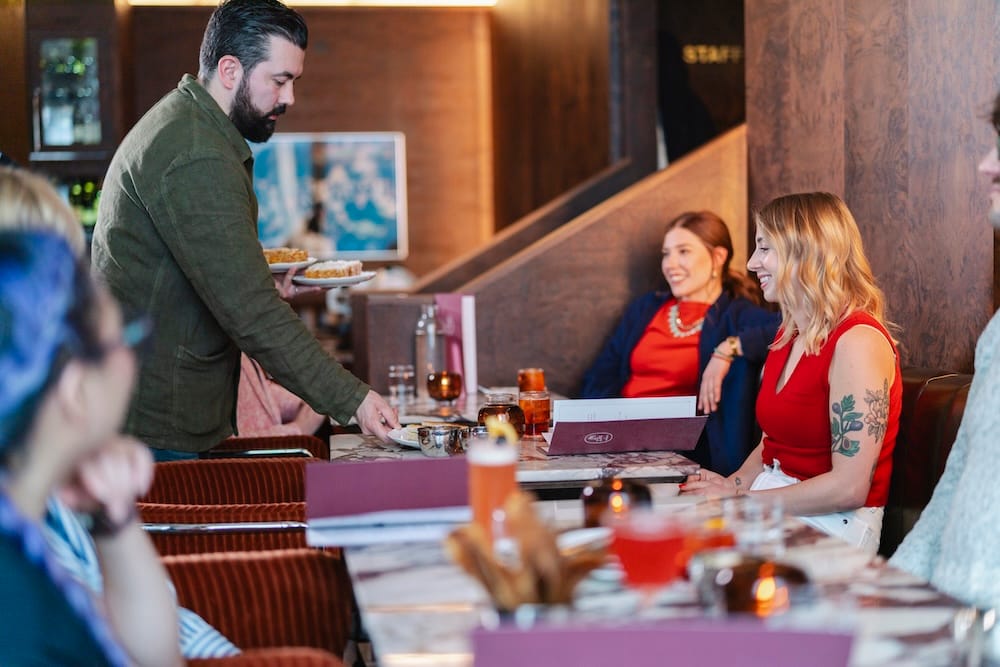 Edmonton restaurant dining and local food scene in Alberta, Canada Diners gather around restaurant tables in Edmonton, Alberta, Canada, as a server delivers plates of food in a warm wood-panelled dining room with drinks and shared dishes on the tables.