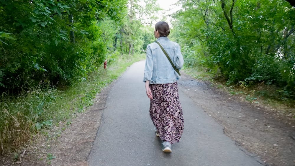 Audrey Bergner walking along a paved trail in Edmonton’s river valley in Alberta, Canada, surrounded by dense green trees and vegetation on both sides of the path.