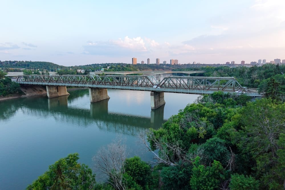 Elevated view across Edmonton’s North Saskatchewan River valley in Alberta, Canada, with a steel truss bridge spanning the water, dense green trees along the banks and the city skyline in the distance.