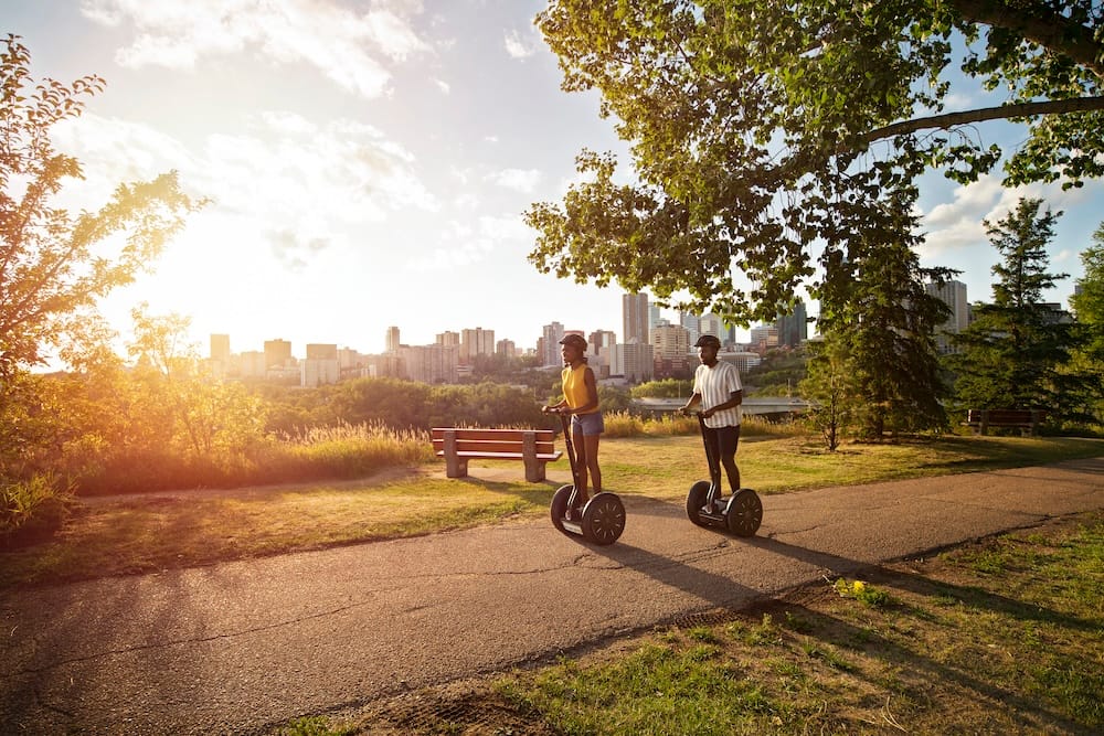 Edmonton Segway tour through the river valley with downtown skyline views in Alberta, Canada Two people ride Segways along a paved path in Edmonton’s river valley, Alberta, Canada, with trees and parkland in the foreground and the downtown skyline glowing in the distance.
