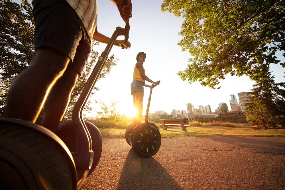 Segway sightseeing tour in Edmonton’s river valley with downtown skyline views, Alberta, Canada Two people ride Segways on a paved trail in Edmonton’s river valley, Alberta, Canada, with the low sun behind them and the downtown skyline visible beyond the trees and parkland.
