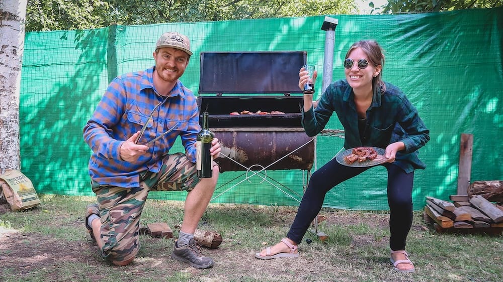 El Bolsón campo asado with Samuel Jeffery and Audrey Bergner grilling outdoors in Argentine Patagonia, holding wine and a plate of grilled meat beside a rustic parrilla during Project 23 Argentina fieldwork.