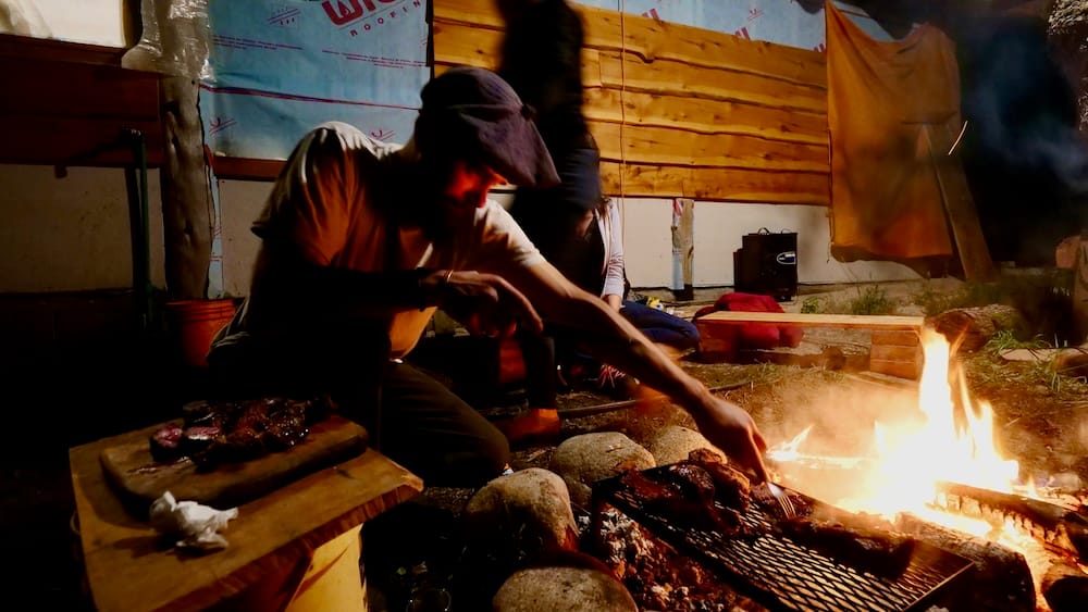 Evening Asado by Firelight — El Bolsón, Río Negro, Patagonia, Argentina | Patagonian Woods Campfire Scene A person tends meat over an open wood fire at night during an evening asado in the Patagonian woods near El Bolsón, Río Negro, Argentine Patagonia, with a rustic wooden shelter and firelight glowing in the background.
