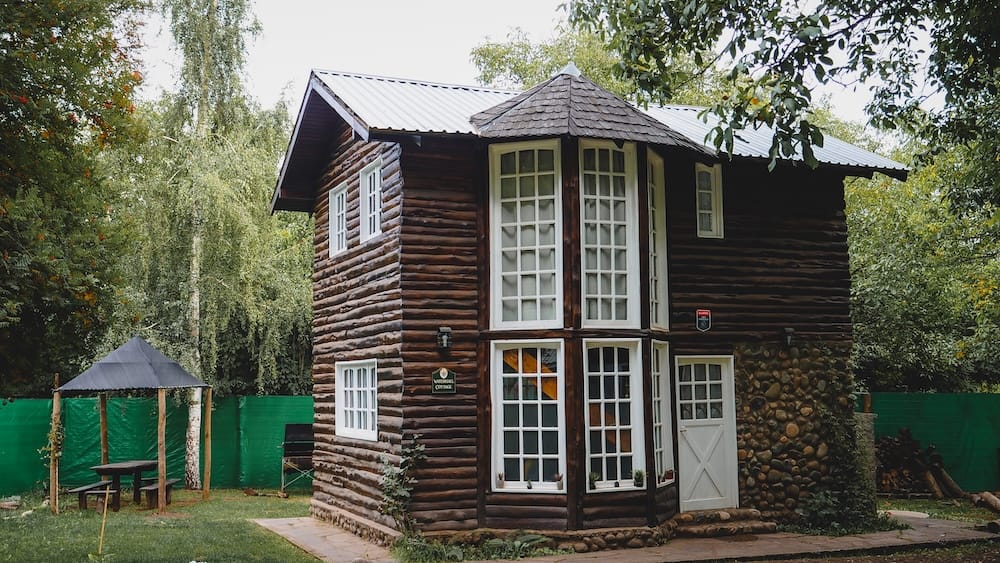 Rustic two-storey log cottage surrounded by trees and garden space in El Bolsón, Río Negro, Patagonia, Argentina, with large windows, timber walls and an outdoor seating area.