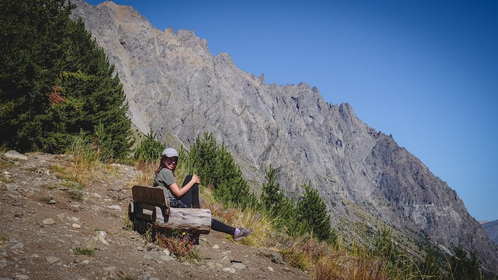 Audrey Bergner descansa en un banco rústico de troncos sobre una ladera rocosa cerca de El Bolsón, Río Negro, Patagonia, Argentina, con pinos y montañas escarpadas detrás bajo un cielo azul despejado.