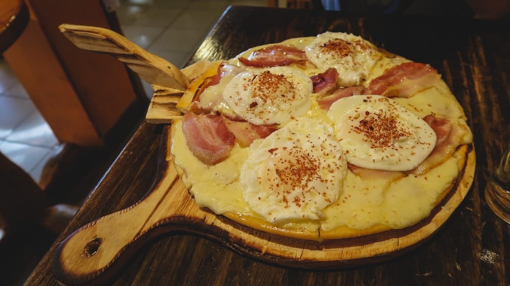 El Bolsón Patagonia bacon and egg pizza showcasing hearty comfort food in Río Negro Argentina dining scene El Bolsón, Río Negro Patagonia — hearty bacon and egg pizza topped with melted cheese and perfectly cooked eggs, served on a wooden board, highlighting the indulgent comfort food and backpacker-friendly dining scene in this Andean town.