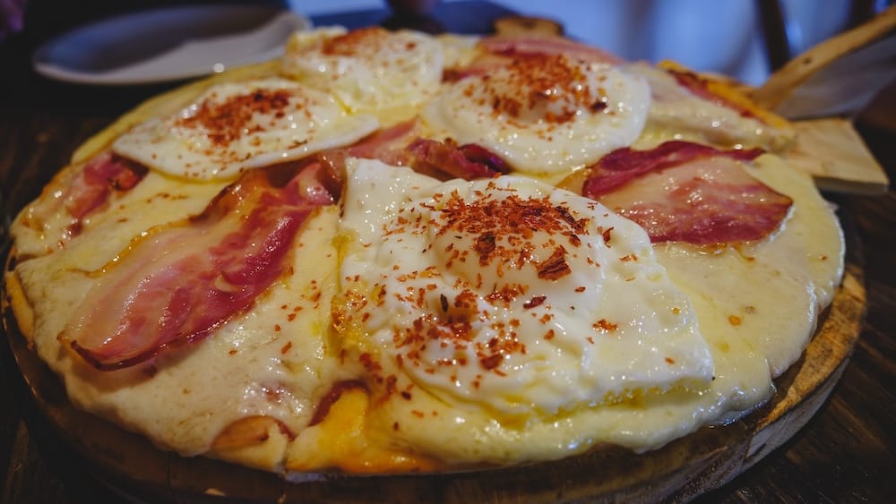 El Bolson Patagonia Argentina pizza topped with fried eggs bacon and melted cheese on rustic crust showcasing hearty mountain town comfort food built for cold Patagonian climate with rich toppings and indulgent local style
