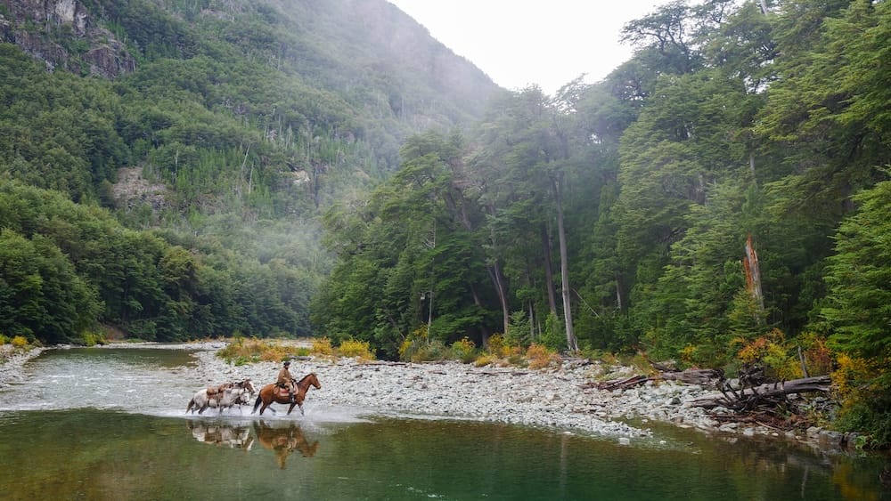 Un guía gaucho lleva caballos a través de un río poco profundo cerca de El Bolsón, Río Negro, en la Patagonia argentina, con montañas boscosas envueltas en niebla y una costa rocosa alrededor del cruce.