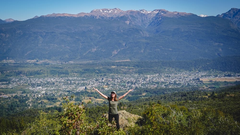 Audrey Bergner Overlooking El Bolsón, Río Negro, Patagonia, Argentina Audrey Bergner stands on a rocky overlook with arms raised above the town of El Bolsón, Río Negro, in Argentine Patagonia, with the valley and snow-dusted mountains spread out behind her under clear blue sky.