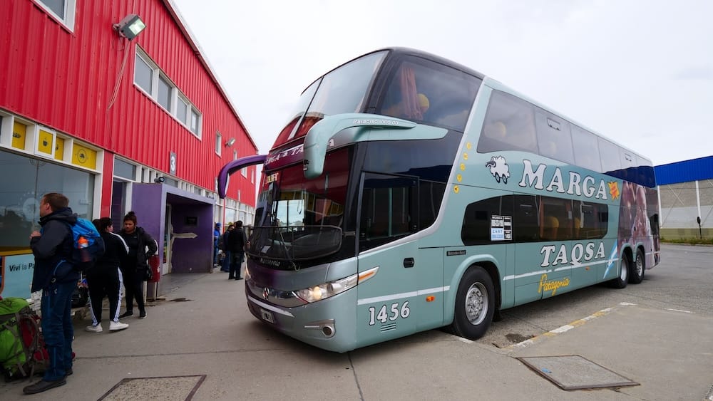 Marga Taqsa Coach at El Calafate Bus Terminal in Santa Cruz, Patagonia, Argentina A Marga Taqsa double-decker coach waits beside the red El Calafate bus terminal in Santa Cruz, Patagonia, Argentina, while passengers stand with backpacks and luggage near the boarding area.