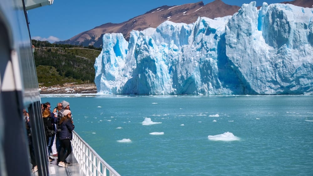 Passengers bundled in jackets on a glacier boat tour in El Calafate, standing beside towering blue ice walls while exposed to cold Patagonian wind over open water, where gusts intensify conditions and lower perceived temperatures dramatically.
