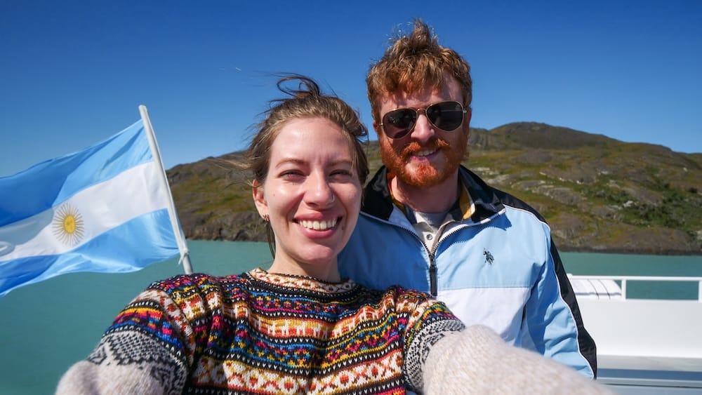 Samuel Jeffery and Audrey Bergner on a boat tour near El Calafate, wind visibly whipping their hair and clothing beside an Argentine flag, demonstrating how Patagonia’s strong gusts impact even sheltered glacier excursions on the water.

