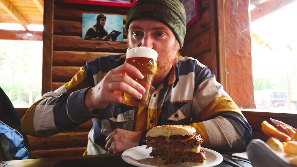 Samuel Jeffery enjoying a large burger and beer indoors in El Calafate after a wind-exposed hike, highlighting the need for high-calorie meals and warm shelter to recover from Patagonia’s physically draining weather conditions.
