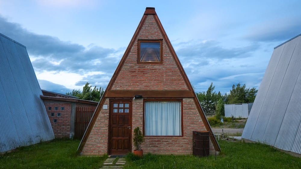 A-Frame Brick Cabin Accommodation in El Calafate, Santa Cruz, Patagonia, Argentina A brick A-frame cabin with a small upper window and curtained lower window stands under an overcast evening sky in El Calafate, Santa Cruz Province, Argentine Patagonia, flanked by corrugated metal outbuildings and grass.