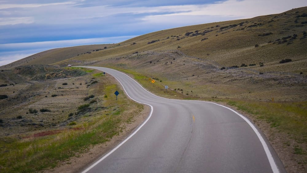 Route Between Río Gallegos and El Calafate — Santa Cruz, Patagonia, Argentina | Steppe Highway Landscape A quiet paved highway winds through rolling, windswept steppe grassland on the route between Río Gallegos and El Calafate in Santa Cruz, Argentine Patagonia, with low hills and a cloudy sky stretching to the horizon.
