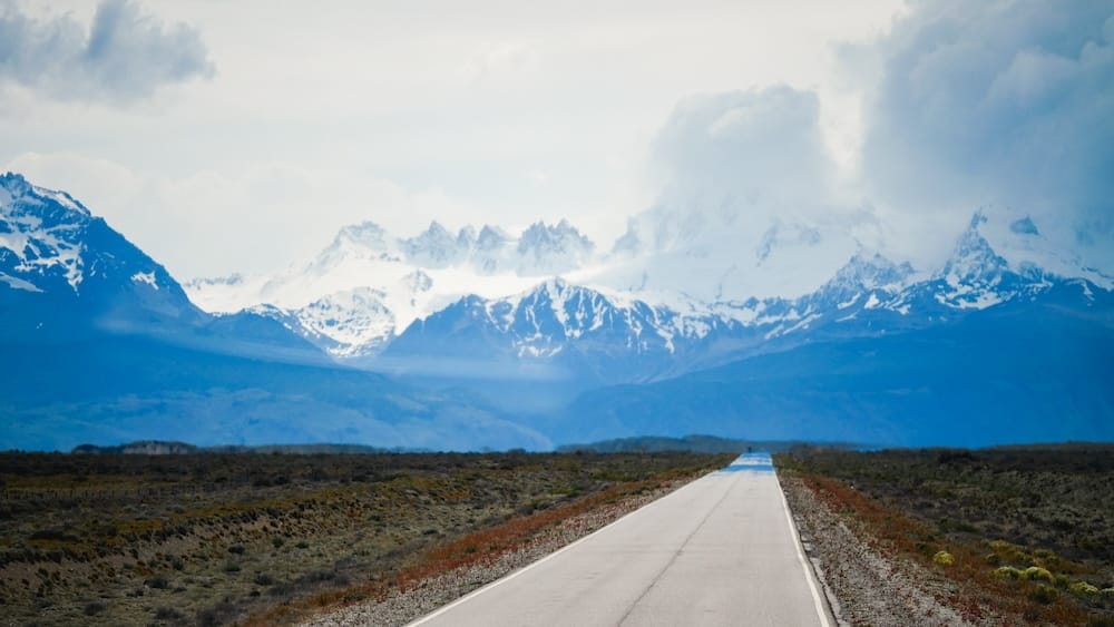 Road from El Calafate to El Chaltén with snowy Andean peaks in Santa Cruz, Patagonia, Argentina A straight paved road stretches across the open Patagonian steppe toward snow-covered Andean peaks beneath heavy clouds on the bus route from El Calafate to El Chaltén in Santa Cruz, Argentina.