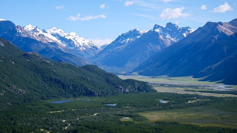 El Chaltén, Argentina view from Mirador de las Águilas overlooking a wide Patagonian valley with forested hills, braided river flats, and distant snow-capped mountains, showing the expansive, open feel of this easy viewpoint hike beyond town.

