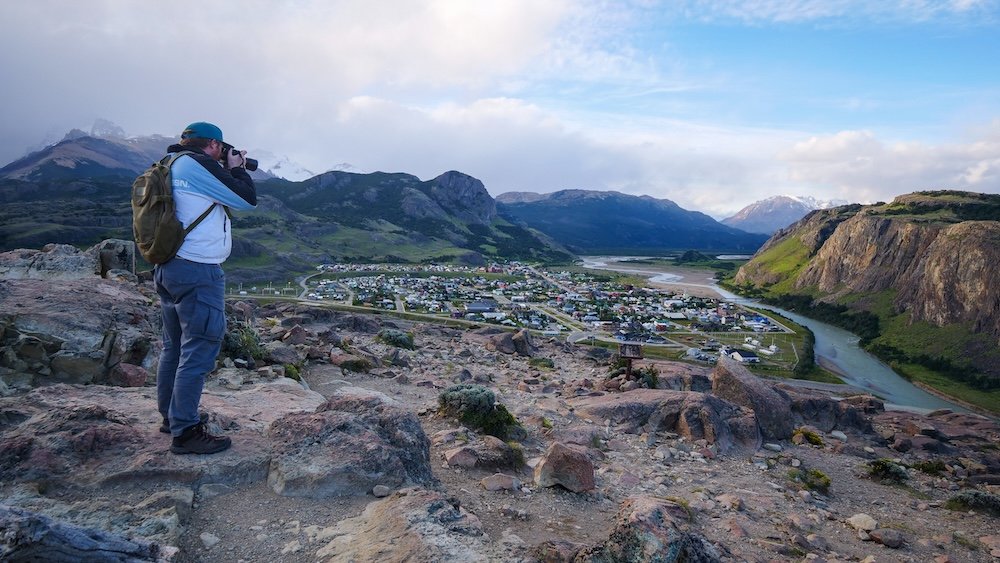 El Chaltén, Argentina seen from a high viewpoint as Nomadic Samuel photographs the town below, with winding rivers, rugged hills, and Patagonian mountains surrounding the village—showcasing the dramatic perspectives you can reach on short hikes near town.