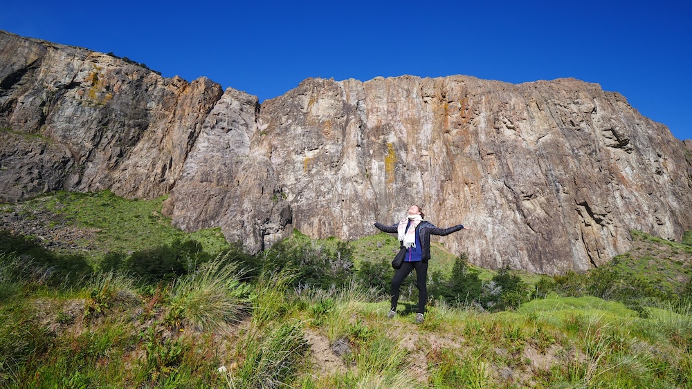 El Chaltén cliffs towering over Patagonia as Audrey Bergner stands with arms raised, thrilled to have arrived in Argentina’s hiking capital, capturing the excitement of the first arrival moment after traveling from El Calafate.