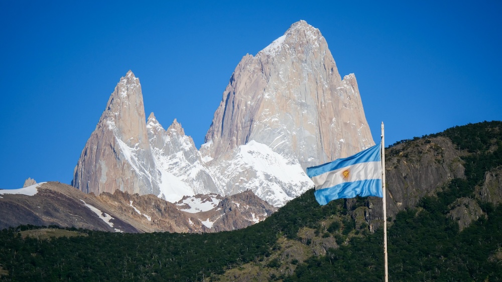 Fitz Roy towering above El Chaltén, Patagonia, Argentina, with a blue-and-white Argentine flag flapping in fierce mountain winds, snowcapped granite peaks glowing under a clear sky in Los Glaciares National Park.