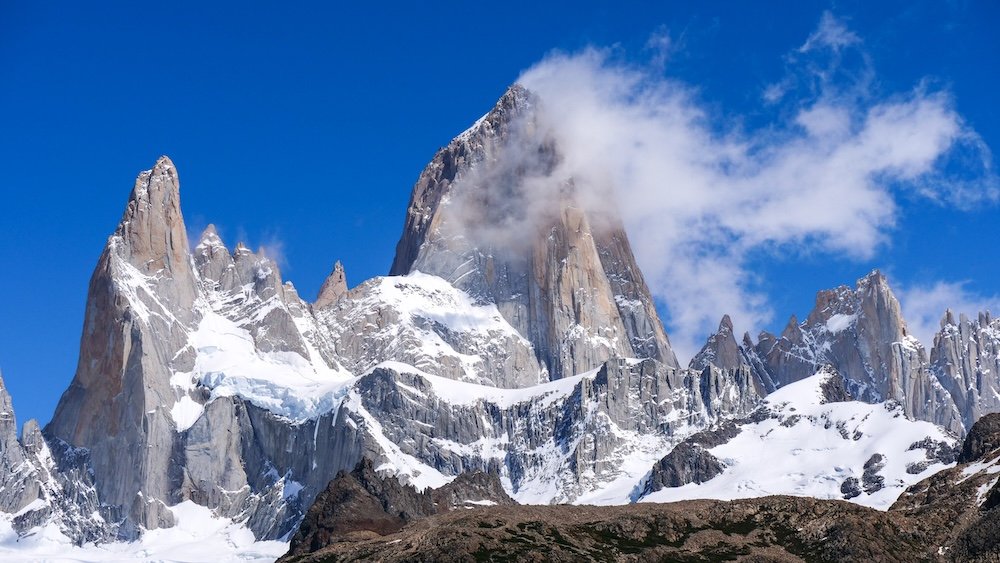 Mount Fitz Roy towering above El Chaltén, Patagonia, with jagged granite spires, snow-draped faces, and clouds curling around the peaks—an iconic sight that rewards hikers who stay long enough to catch clear weather windows.
