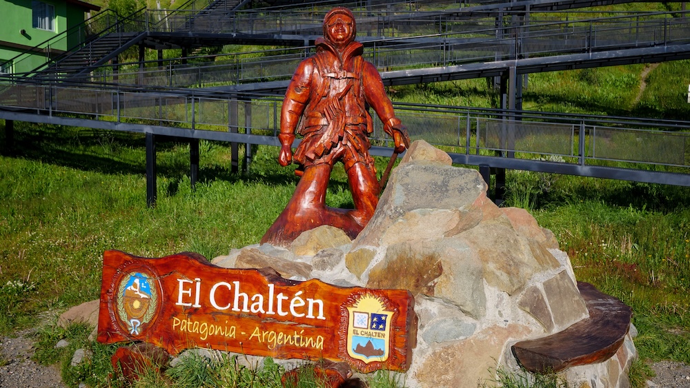 El Chaltén hiker monument in Patagonia, Argentina, featuring a carved wooden mountaineer statue standing atop stonework with a colorful town sign, marking the trailhead gateway to Argentina’s famous hiking capital.