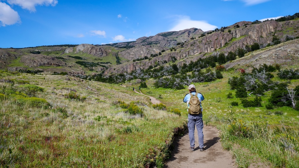 Nomadic Samuel hiking the Laguna Torre trail in El Chaltén, Argentina, enjoying sunny December weather surrounded by lush green valleys and dramatic mountain cliffs, camera in hand capturing the beauty of Patagonia’s rugged landscapes.