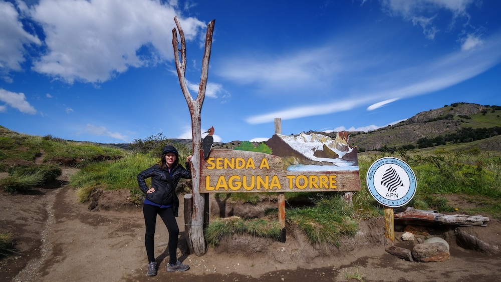 El Chaltén Patagonia Laguna Torre trailhead entrance sign marking the start of the famous hiking route in Los Glaciares National Park, where Audrey Bergner stands beside the carved wooden marker used by trekkers to navigate the trail without relying on GPS.