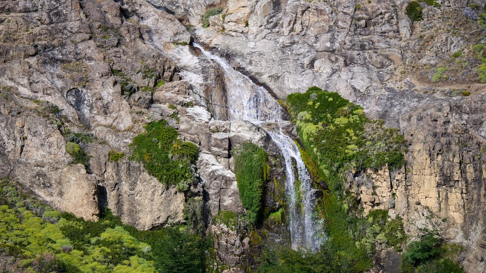A waterfall cascading down steep granite cliffs along the Laguna Torre hike in El Chaltén, Patagonia, Argentina, with green moss-covered rock faces and rugged mountain scenery inside Los Glaciares National Park.
