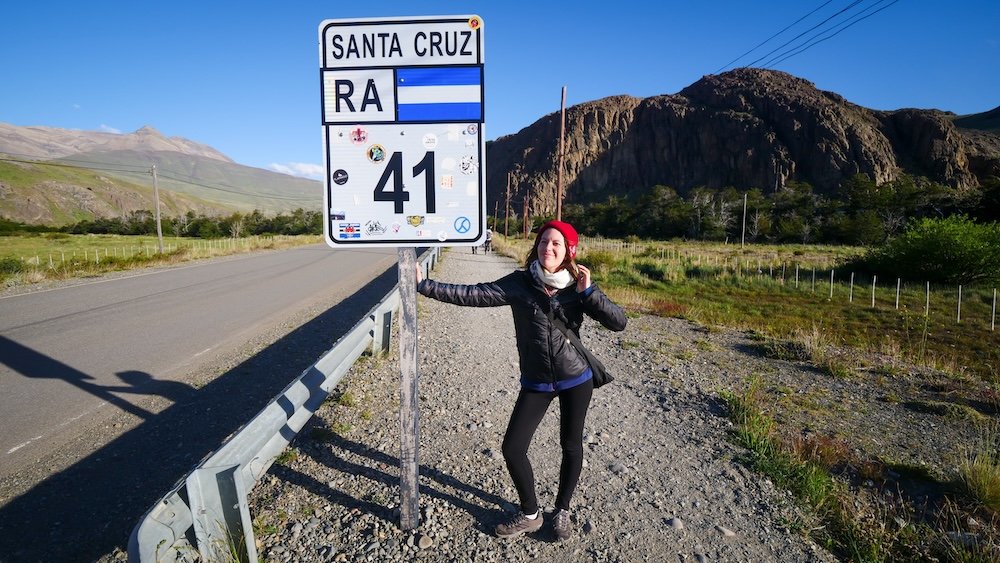 Audrey Bergner arriving in El Chaltén on day one, Route 41 sign in Santa Cruz Province, Patagonia Audrey Bergner arriving in El Chaltén, Patagonia, on day one of our trip, standing beside the Route 41 sign in Santa Cruz Province, Argentina, with open road, dramatic rocky hills, and clear blue skies setting the tone for a hiking-focused adventure.